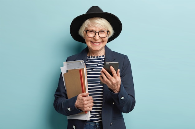 Femme d'affaires souriante âgée réussie vérifie les informations de données sur le téléphone mobile, tient le bloc-notes avec des documents, revient d'une conférence importante, vêtue de vêtements élégants, effectue le paiement en ligne