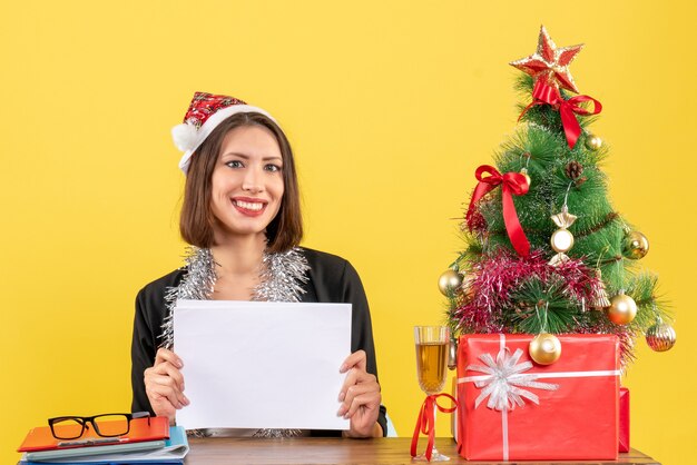 Femme d'affaires souriante en costume avec chapeau de père Noël et décorations de nouvel an travaillant seul tenant des documents et assis à une table avec un arbre de Noël dessus dans le bureau