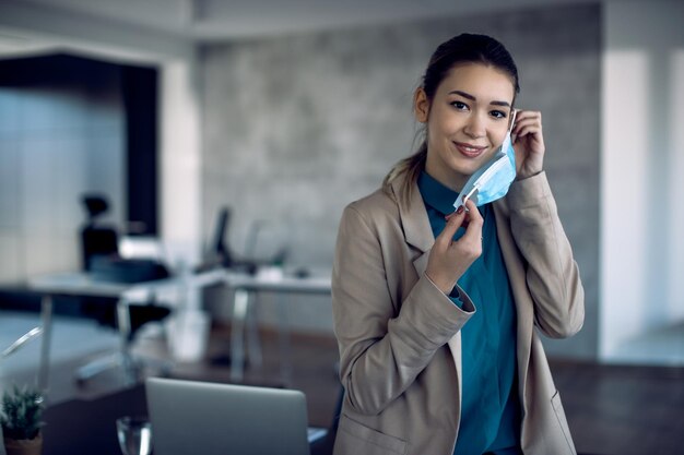 Femme d'affaires souriante mettant un masque protecteur au bureau et regardant la caméra