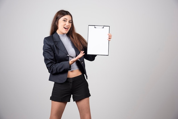 Une femme d'affaires souriante montrant un presse-papiers isolé sur un mur gris.