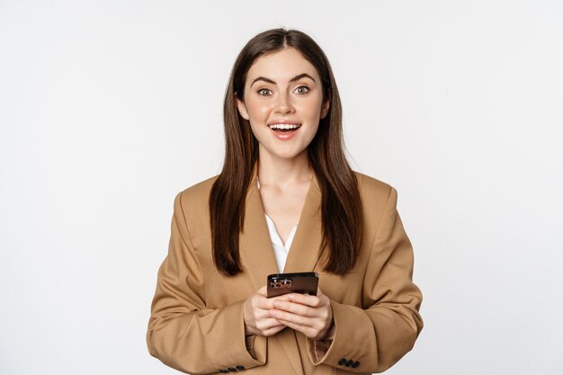 Femme d'affaires souriante utilisant une application pour smartphone sur un téléphone portable debout sur fond blanc