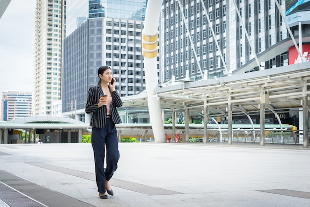 Femme d&#39;affaires utilisant un téléphone avec du café à la main, marchant dans la rue avec des immeubles de bureaux en arrière-plan