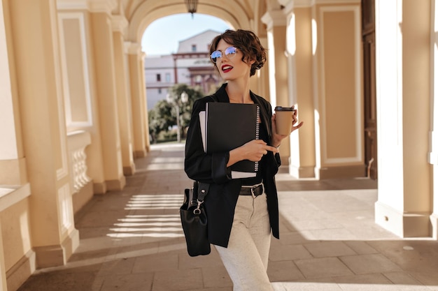 Femme d'affaires en veste sombre et pantalon blanc posant avec une tasse de café à l'extérieur