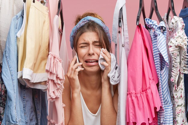 Femme affligée qui panique sans rien avoir à porter, regarde à travers un étagère de vêtements, parle au téléphone intelligent, pleure d'insatisfaction.