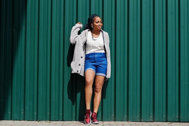 Photo gratuite femme africaine avec des cheveux dreads en short jeans posé contre un mur d'acier vert