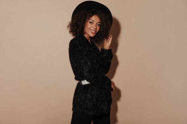 Femme africaine avec une courte coiffure bouclée en chapeau noir et des vêtements brillants élégants souriant et regardant la caméra sur un mur beige.