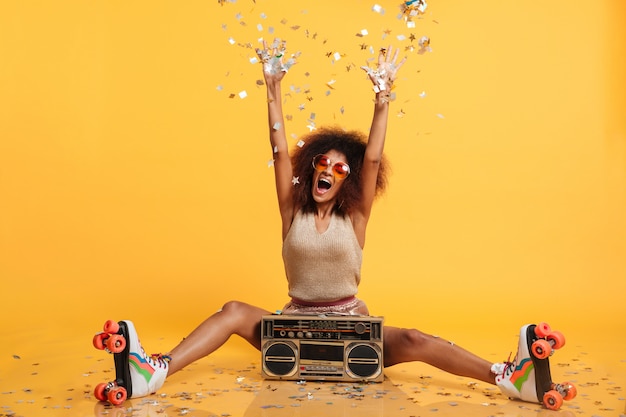 Femme africaine disko émotionnelle en vêtements rétro et scates à rouleaux jetant des confettis assis avec boombox