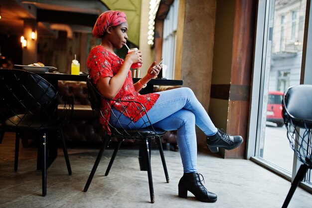 Femme africaine élégante en chemise rouge et chapeau posé café intérieur et boire de la limonade aux fraises avec téléphone à portée de main