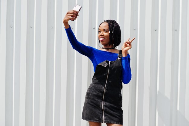 Femme africaine en jupe salopette en jean et robe bleue posée contre un mur en acier blanc et faisant selfie au téléphone