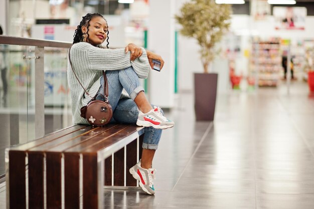 Femme africaine posée en pull et jeans posés au centre commercial assis sur un banc avec son téléphone portable