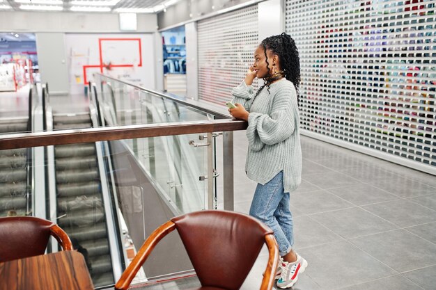 Femme africaine posée en pull et jeans posés au centre commercial assis à table avec téléphone portable