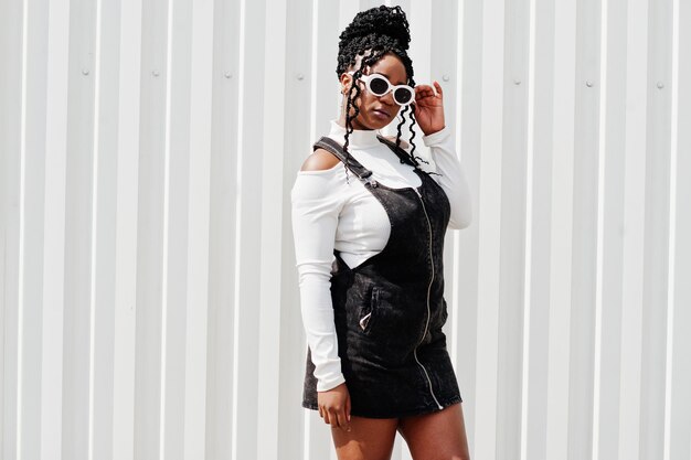 Femme africaine en salopette en jean jupe lunettes de soleil blanches posées contre un mur en acier blanc