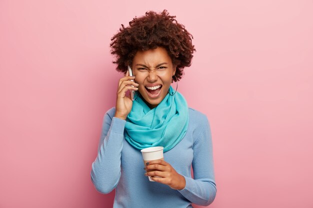 Une femme afro agacée se dispute via un smartphone, crie fort de frustration, tient une tasse de café à emporter, porte un pull bleu avec un foulard