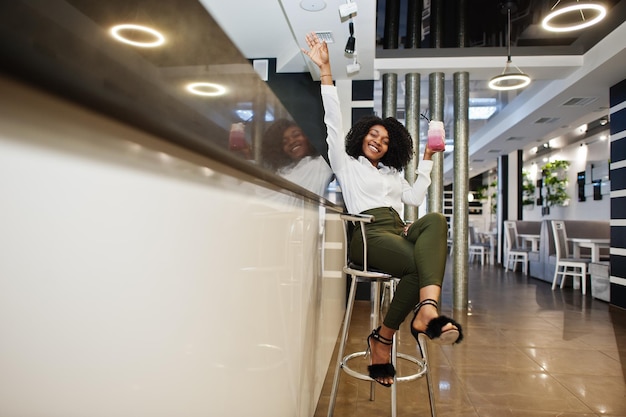 Une femme afro-américaine d'affaires pure aux cheveux afro porte un chemisier blanc et un pantalon vert assis sur le comptoir du bar et boit un cocktail rose