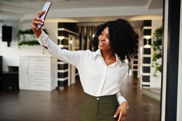 Une femme afro-américaine d'affaires pure avec des cheveux afro porte un chemisier blanc et un pantalon vert posés dans un café avec un téléphone portable faisant du selfie