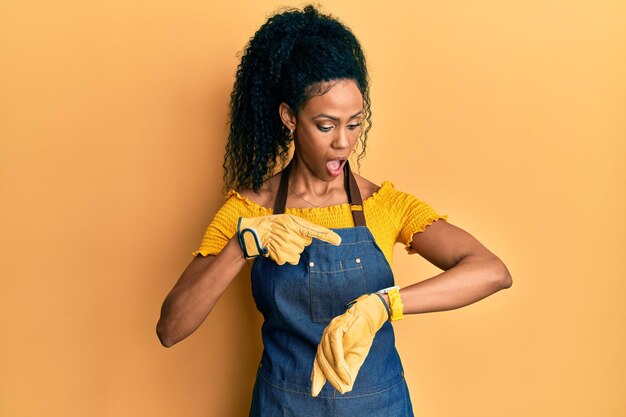 Femme afro-américaine d'âge moyen portant un tablier professionnel pressé pointant vers l'impatience de l'heure contrariée et en colère pour le retard de l'échéance