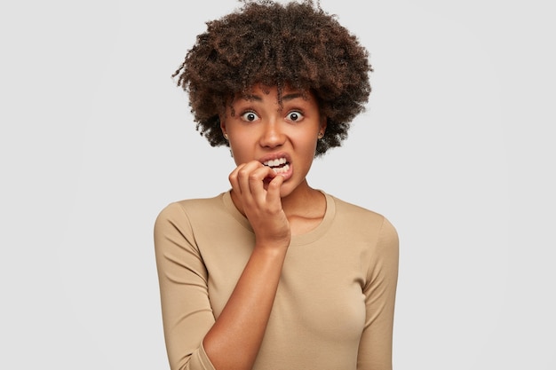 Photo gratuite une femme afro-américaine anxieuse et nerveuse se mord les ongles avec une expression perplexe, a les cheveux noirs bouclés, habillée avec désinvolture, isolée sur un mur blanc. omg, j'ai peur de ça! concept d'émotions