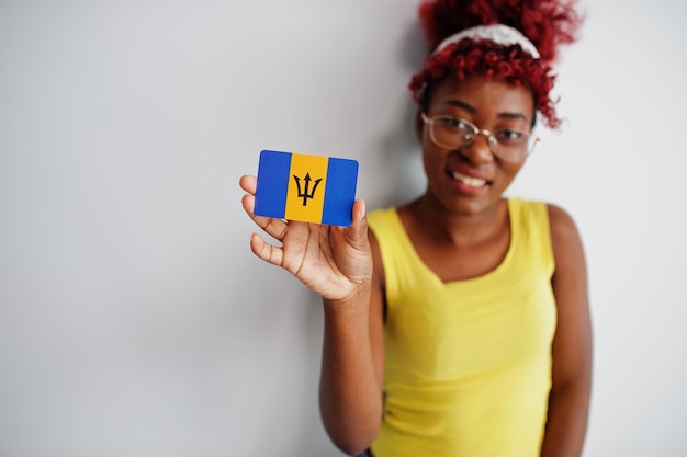 Une femme afro-américaine aux cheveux afro porte un maillot jaune et des lunettes tient le drapeau de la Barbade isolé sur fond blanc