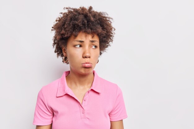 Une femme afro-américaine aux cheveux bouclés bouleversée porte les lèvres et regarde tristement loin a une expression misérable vêtue d'un t-shirt rose décontracté pose contre un mur blanc
