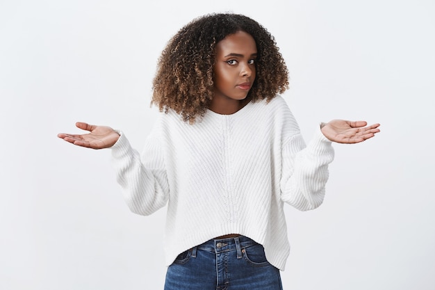 Photo gratuite une femme afro-américaine aux cheveux bouclés inconsciente, ignorante, haussant les épaules, tourne le visage en secouant la tête interrogée, écarte les mains sur le côté, n'a aucune idée, debout douteuse, mur blanc