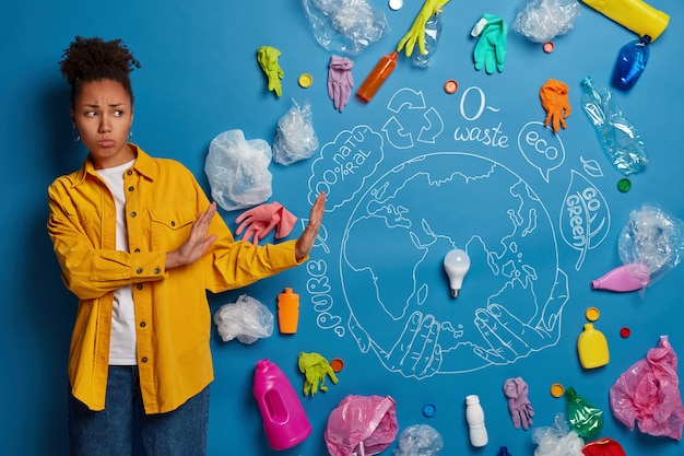 Une femme afro-américaine bouclée mécontente fait un geste d'arrêt, nie avoir utilisé du plastique, regarde tristement les ordures et les déchets, est engagée dans le recyclage des ordures, veut vivre dans un environnement propre.