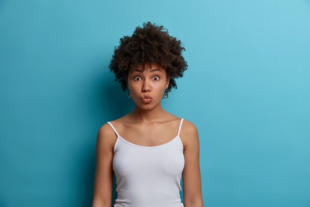 Une femme afro-américaine choquée garde les lèvres arrondies, se sent étonnée, remarque une chute de prix inattendue impressionnante, habillée avec désinvolture, fait une drôle de grimace, isolée sur un mur bleu. Expressions de visage humain