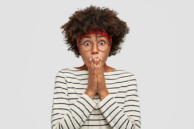 Une Femme Afro-américaine Choquée Regarde Avec Des Yeux écarquillés, Couvre La Bouche Avec Des Paumes, Vêtue De Vêtements Décontractés, Exprime La Surprise, Se Tient Contre Un Mur Blanc. Fille émotive à La Peau Sombre