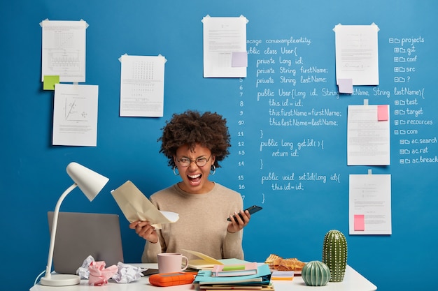 Une femme afro-américaine en colère émotionnelle tient un document papier et un téléphone portable, frustrée par l'échec de la réussite du projet.