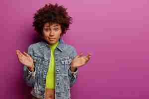Photo gratuite une femme afro-américaine douteuse et désemparée écarte les paumes avec hésitation, ne peut pas prendre de décision, porte une veste et un haut en jean, a un regard perplexe, pose sur un mur violet vibrant, espace de copie de côté