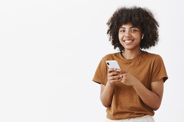 Femme afro-américaine élégante et heureuse moderne à l'aide d'écouteurs sans fil tout en parlant avec des clients tenant un smartphone en prenant des notes ou en choisissant de la musique dans l'appareil