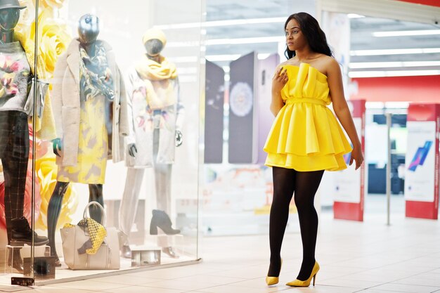 Femme afro-américaine élégante à la robe jaune posée contre des vitrines avec des mannequins au centre commercial