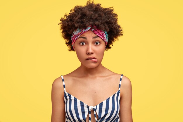 Une femme afro-américaine embarrassée porte les lèvres, regarde avec perplexité, entend quelque chose de pas clair, vêtue d'un haut marin décontracté, isolée sur un mur jaune. Personnes et expressions faciales