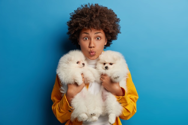 Femme afro-américaine embrasse deux chiots blancs, garde les lèvres pliées comme veut embrasser de beaux animaux de compagnie, passe le week-end avec ses meilleurs amis