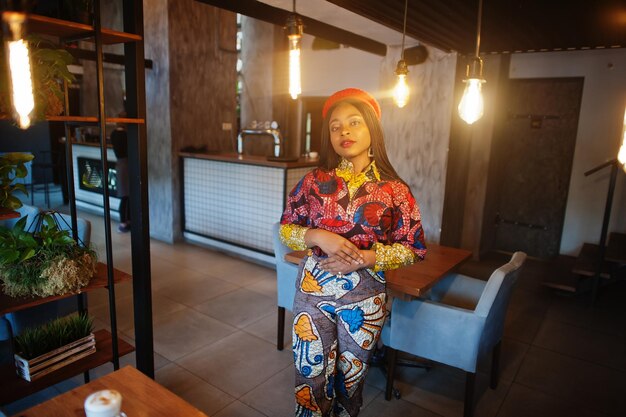 Femme afro-américaine enthousiaste en tenue colorée à la mode avec béret rouge se relaxant dans un café confortable avec de nombreuses lampes