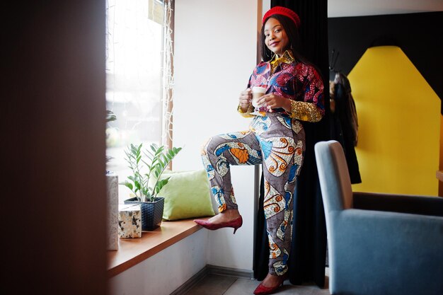 Femme afro-américaine enthousiaste en tenue colorée à la mode avec béret rouge se relaxant dans un café confortable avec une tasse de latte chaud dans les mains