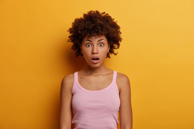 Une femme afro-américaine étonnée se rend compte du mensonge d'une personne proche, regarde avec une expression de visage choquée, ouvre la bouche, se tient dans la stupeur contre le mur jaune. Réaction humaine et émotions