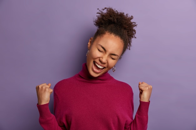 Photo gratuite une femme afro-américaine euphorique réussie célèbre une nouvelle incroyable, chanceuse de gagner beaucoup d'argent, triomphe comme rêve devenu réalité, incline la tête, vêtue d'un col roulé décontracté, isolée sur un mur violet