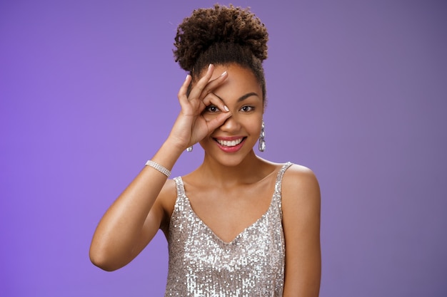 Une femme afro-américaine féminine élégante et confiante en robe argentée à la mode montre un geste d'accord sur l'œil regardant vers l'avant avec plaisir souriant, sûr de lui comme la perfection, sur fond bleu.