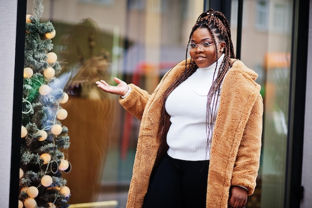 Femme afro-américaine glamour en manteau de fourrure chaud pose dans la rue près de l'arbre de noël