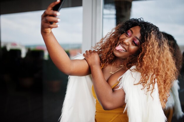 Femme afro-américaine glamour en robe jaune et cape en laine blanche faisant selfie au téléphone