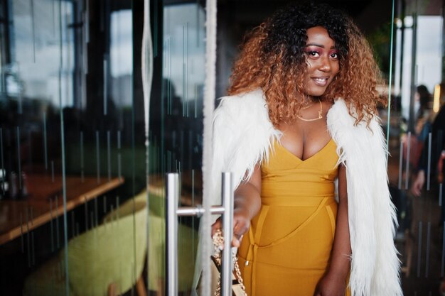 Femme afro-américaine glamour en robe jaune et cape en laine blanche avec sac à main posé à l'entrée de la porte vitrée du restaurant