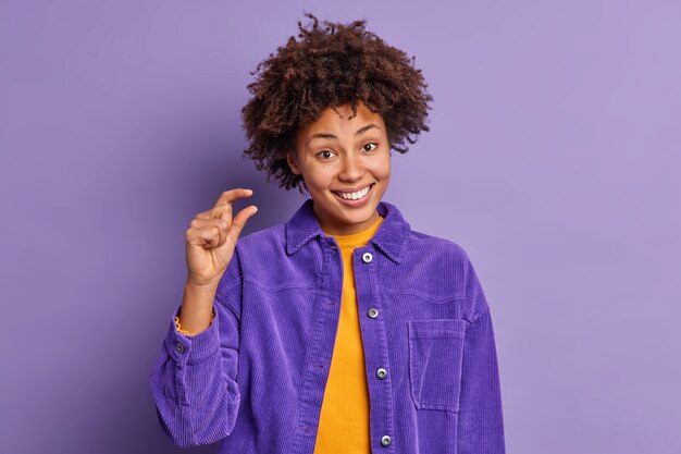 Une femme afro-américaine heureuse aux cheveux bouclés montre des stands de petite taille joyeux montre une petite chose ou un objet vêtu d'une veste de velours pose contre un mur violet. Concept de langage corporel