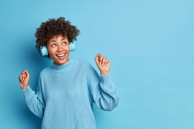 Photo gratuite une femme afro-américaine insouciante positive danse insouciante garde les bras lever les sourires s'amuse largement aime la musique préférée porte des écouteurs sans fil habillés en pull