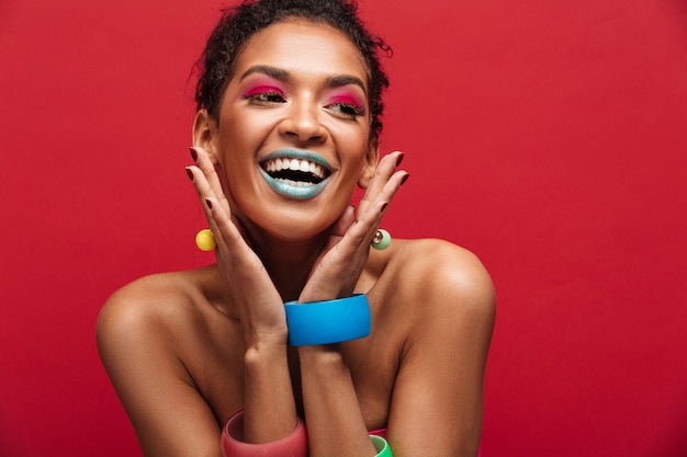 Femme afro-américaine joyeuse multicolore avec maquillage mode souriant et regardant de côté, isolé sur mur rouge