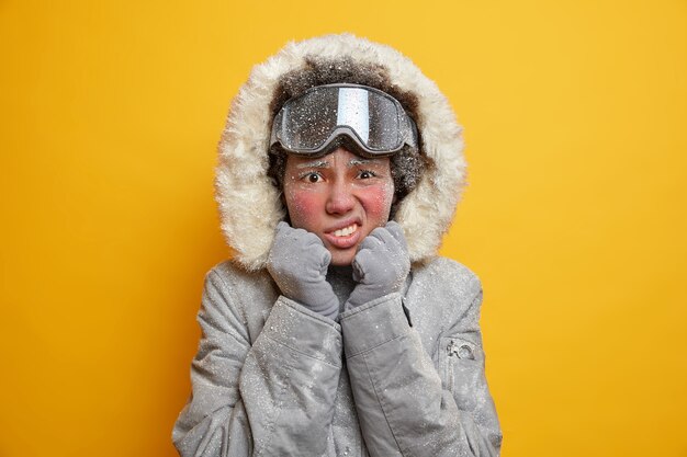 Une femme afro-américaine mécontente a le visage couvert de glace serre les dents du froid regarde malheureusement porte une veste et des lunettes de ski profite des vacances d'été tremble par temps glacial