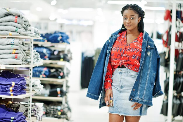 Femme afro-américaine à la mode en chemise rouge et jupe en jean avec veste posée au magasin de vêtements Il est temps de faire du shopping