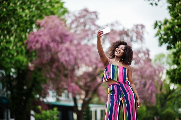 Femme afro-américaine à la mode en combinaison rayée rose posée dans la rue de la floraison printanière et faisant selfie par téléphone mobile