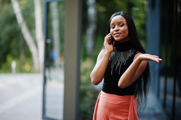 Femme afro-américaine à la mode en pantalon pêche et chemisier noir parler au téléphone en plein air