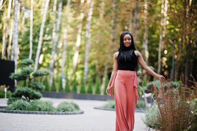 Femme afro-américaine à la mode en pantalon pêche et chemisier noir pose en plein air