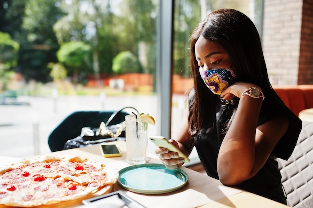 Photo gratuite une femme afro-américaine à la mode porte un masque facial fait à la main assis au restaurant regarde son téléphone portable tout en mangeant de la pizza nouvelle vie normale